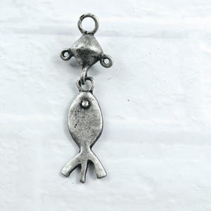 May include: A silver-toned metal charm featuring a fish-shaped pendant suspended from a triangular connector piece. The charm has a distressed, matte finish with a loop at the top for attachment, showcasing a simple, flat, and elongated design.