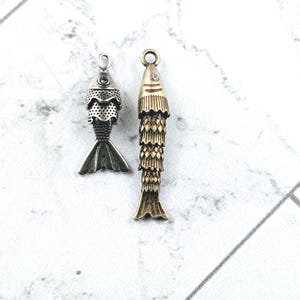 May include: Two fish-shaped pendants. One is silver-toned with a textured body and tail, and a small gem. The other is gold-toned, with a scaled body and tail, and a small gem. Both have a loop for hanging.