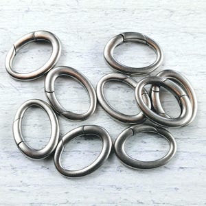 May include: A group of eight silver oval-shaped jump rings. The rings are all the same size and are arranged in a loose pile.