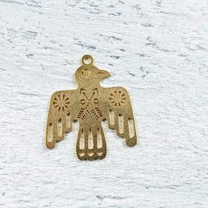 May include: A gold metal charm in the shape of a stylized bird with geometric patterns. The bird has a long beak and spread wings with intricate details.