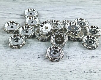 May include: A group of silver-toned metal bead caps with clear rhinestones. The bead caps are round and have a raised edge. They are arranged in a random pattern on a white surface.