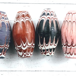May include: Four decorative, elongated glass beads with a swirled pattern. The beads are in shades of purple, brown, black, and pink, with white and red accents. The beads have a glossy finish and are arranged side by side.