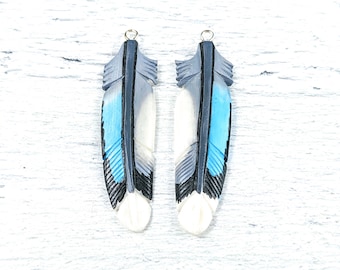 May include: Two feather-shaped pendants. Each pendant features a detailed design with shades of blue, gray, black, and white. The feathers have a carved appearance and are topped with a small gold-colored loop for hanging.