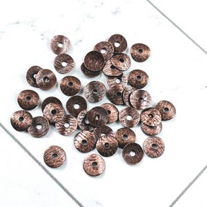 May include: A collection of small, round, copper-colored metal beads with a textured surface and a central hole. The beads are scattered on a white surface, ideal for jewelry making or craft projects.