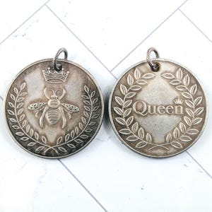 May include: Two silver-toned pendants. One features a bee with a crown above and a laurel wreath. The other pendant has the word "Queen" with a crown above and a laurel wreath. Both pendants have a loop at the top.