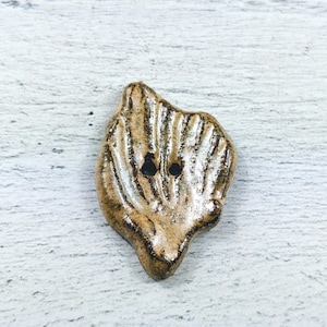 May include: A ceramic button with a textured, leaf-like design in shades of brown and gold. The button has two black holes for sewing.
