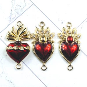 Puede incluir: Tres colgantes en forma de corazón con temática religiosa. Cada colgante presenta un vibrante corazón rojo, acentuado con metal dorado y brillantes cristales transparentes. Los colgantes tienen un lazo en la parte superior e inferior para sujetarlos a la joyería.