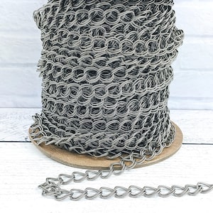 Puede incluir: Un carrete de cadena plateada con un patrón de eslabones en forma de corazón. La cadena está enrollada en un carrete de madera y una longitud de cadena se coloca sobre una superficie blanca.
