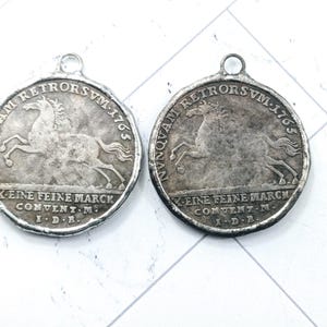 May include: Two antique silver-toned pendants with a circular design. Each pendant features a horse and text, including "NUNQUAM RETRORSVM-1765" and "EINE FEINE MARCK CONVENT M I.D.B.". The pendants have a loop at the top for hanging.