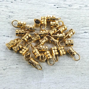 May include: A pile of gold-toned metal crimp beads with a decorative pattern. The beads are designed to be used for jewelry making and other crafts.