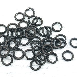 May include: A pile of black metal jump rings. The rings are all the same size and are made of a twisted metal wire.