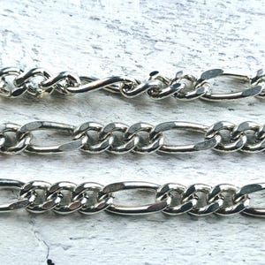 May include: Three silver chains with a curb link pattern. The chains are layered on top of each other, with the top chain being the thinnest and the bottom chain being the thickest.