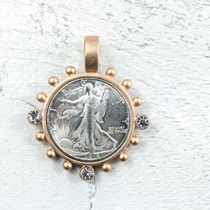 May include: A gold pendant with a silver coin at its centre, depicting a woman walking towards the sun. The coin is framed in gold, with gold beads and clear crystals. It has a loop for a chain.