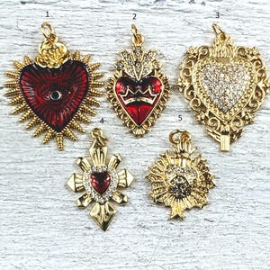 May include: Five gold-tone pendants featuring red and white enamel hearts. The pendants are shaped like a heart, a cross, and a sunburst. All are decorated with rhinestones.