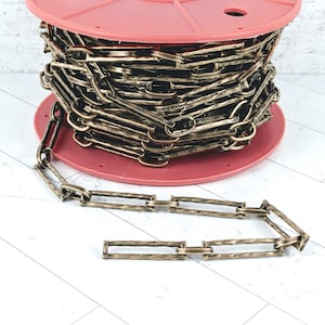 May include: A long, continuous metal chain with rectangular links featuring a textured, antique bronze finish. The chain is neatly wound around a round, coral-colored plastic spool, with a portion of the chain extending outward in front.