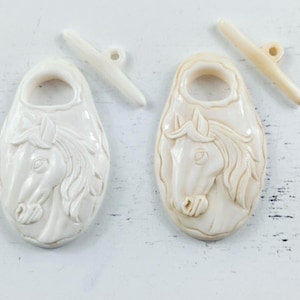 May include: Two off-white toggle buttons, each featuring an oval shape with a raised relief carving of a horse head. Each button includes a separate, matching bar-shaped toggle piece with a single hole, designed for fastening through a loop.