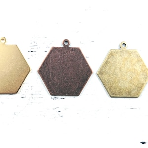 May include: Three metal hexagon charms in gold, bronze, and brass tones. Each charm has a small loop at the top for attaching to jewellery or other crafts. The charms are displayed on a white surface.