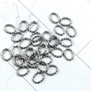 Puede incluir: Un conjunto de pequeños anillos ovalados de color plateado con un diseño de cuerda trenzada. Estos hallazgos de joyería están dispersos sobre una superficie blanca con un sutil patrón gris claro. Ideal para la creación de joyas.