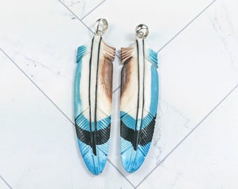 May include: Two feather-shaped pendants with silver-colored bails. The pendants are carved with a detailed feather design. The feathers are painted with shades of blue, white, brown, and black. The pendants are displayed on a white surface.