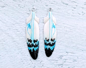 May include: A pair of white feather earrings with blue and black painted designs. The feathers are long and narrow, with a loop at the top for hanging.