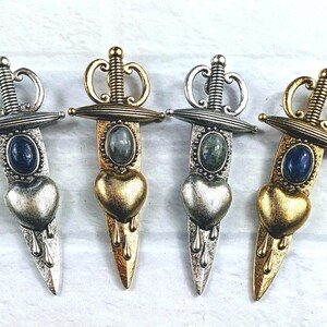May include: Four silver and gold dagger-shaped pendants with heart-shaped charms and gemstone accents. The pendants feature a detailed design with a crown and a sword.