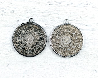 May include: Two silver zodiac pendants with a circular design featuring the twelve signs of the zodiac and Roman numerals. The pendants are on a white wooden surface.