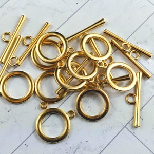 以下が含まれることがあります： 金色の金属製のトグル留め具とジャンプリングのセット。留め具は棒状で、両端にリングが付いています。ジャンプリングは丸い形をしています。