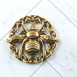 May include: A gold-colored bee brooch with intricate detailing. The bee is centered within a chain-link oval frame. The wings and body of the bee are textured, and the brooch has a vintage aesthetic.