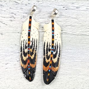 May include: Two carved feather pendants featuring an off-white base with black, orange, and blue painted details. Each feather has a central black spine with colorful dots and a silver-toned metal bail at the top for hanging as a necklace.