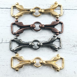 May include: Four metal buckles in gold, bronze, and silver tones. Each buckle features a decorative design with a central ring and a rectangular loop. The buckles are arranged on a white wooden surface.