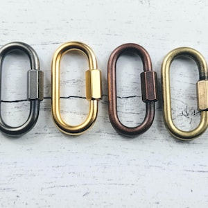 May include: Four oval metal carabiner clasps in different finishes: dark grey, gold, bronze, and brass. Each clasp has a screw-lock mechanism. The clasps are shown on a light-coloured, textured surface.