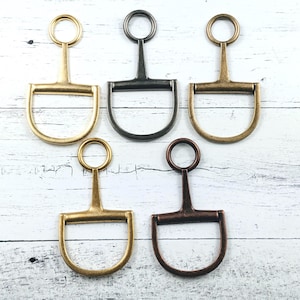 May include: Five metal horse bit charms in various finishes: gold, black, and bronze. Each charm features a D-shaped bit with a central bar and a ring at the top. The charms are arranged on a white wooden surface.