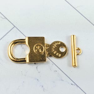 May include: Gold-toned metal clasp with a padlock design and a keyhole. The clasp has a toggle bar with a loop on one end. The padlock has the word "SEEDS" engraved on it.