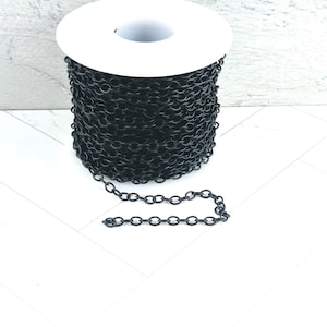 May include: A spool of black chain. The chain is made of small, oval-shaped links. The spool is white and the chain is unspooled at the bottom. The background is white.