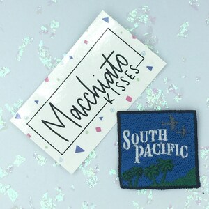 Pode incluir: Um autocolante retangular branco com o texto "Macchiato Kisses" em fonte manuscrita, e um patch quadrado azul com o texto "South Pacific", palmeiras e pássaros.