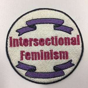 Puede incluir: Un parche bordado blanco con detalles de cinta morada y el texto "Intersectional Feminism" en rojo.