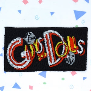 Op de afbeelding: Een zwarte geborduurde patch met het woord "Guy Dolls" in rode, witte en gele letters.