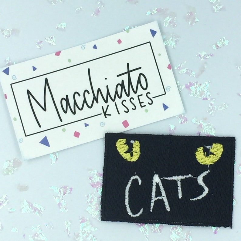 K&ouml;nnte beinhalten: Schwarzer Filz-Patch mit zwei gelben Katzenaugen und dem Wort "CATS" in wei&szlig;er Stickerei.