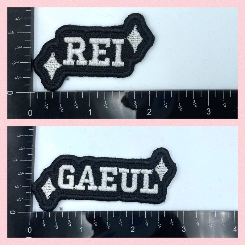 Puede incluir: Dos parches de tela negra con letras bordadas en blanco. El parche superior dice "REI" y el parche inferior dice "GAEUL". Ambos parches tienen una forma de diamante blanco en el extremo.