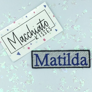 Puede incluir: Un parche de plancha gris con borde negro y texto bordado en azul que dice "Matilda".
