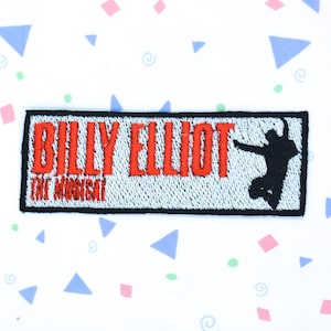 Può includere: Una toppa ricamata bianca e nera con il testo "Billy Elliot The Musical" in lettere rosse. Una silhouette nera di un ballerino è sul lato destro della toppa.