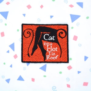 May include: A red embroidered patch featuring a black silhouette of a cat on a roof. The text "Cat on a Hot Tin Roof" is embroidered in white.