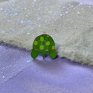 May include: A green enamel pin shaped like a turtle with a hexagonal shell pattern.