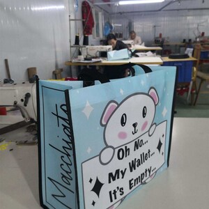 Puede incluir: Una bolsa de tela reutilizable azul y blanca con un oso de dibujos animados sosteniendo un cartel que dice "Oh No... Mi Billetera... Está Vacía..." La bolsa tiene un asa negra y la palabra "Macchiato" escrita en el lateral.