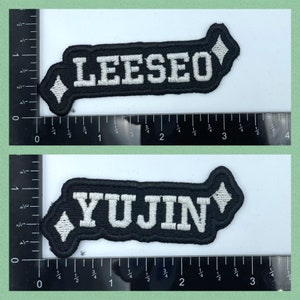 Puede incluir: Dos parches de tela negra con letras blancas. El parche superior dice "LEESEO" y el parche inferior dice "YUJIN".