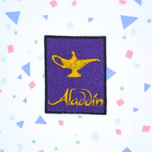 May include: Purple embroidered patch featuring a gold genie lamp and the word "Aladdin" in gold lettering.