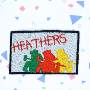 Peut inclure: Un patch brodé blanc avec le mot "HEATHERS" en lettres rouges. Trois figures sont brodées en vert, rouge et jaune.