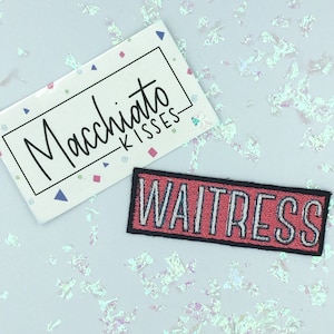 Puede incluir: Parche bordado rosa y negro con la palabra "WAITRESS" en letras blancas.