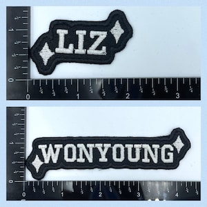 Puede incluir: Dos parches bordados negros con letras blancas. El primer parche dice "LIZ" y tiene forma de diamante. El segundo parche dice "WONYOUNG" y es rectangular.