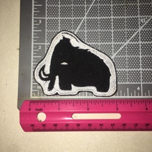 May include: A black embroidered patch in the shape of a woolly mammoth with a white border. The patch is positioned on a cutting mat next to a pink ruler, showing the design measures approximately 3.5 inches in width and 2.5 inches in height.